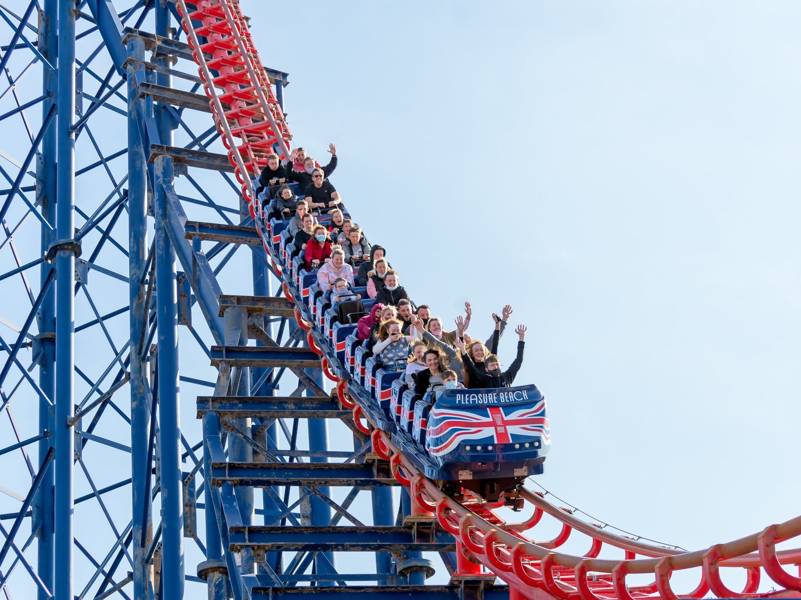 People riding a roller coaster at Blackpool Pleasure Beach, waving their hands as the train descends a steep red and blue track under a clear blue sky.
